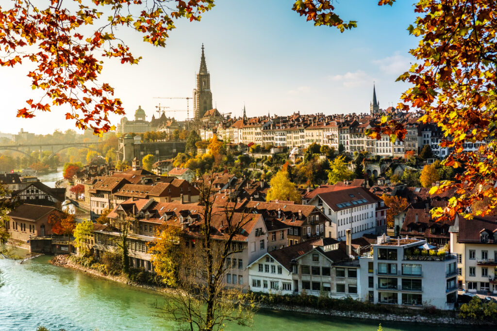 Die Berner Altstadt am Flussufer, im Vordergrund hängen herbstliche Blätter an Ästen, im Hintergrund erkennt man eine Brücke und mehrere Kirchtürme