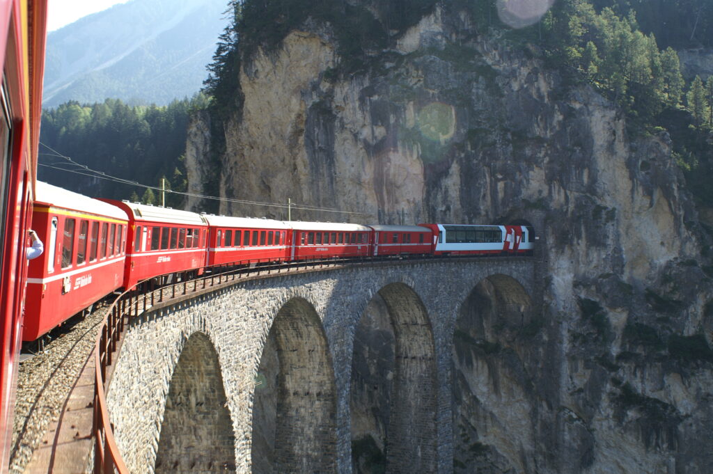 Ein roter Zug der Rhätischen Bahn, der über eine gebogene Steinbrücke fährt und in einem Tunnel im Berg verschwindet, Top Sehenswürdigkeit in der Schweiz