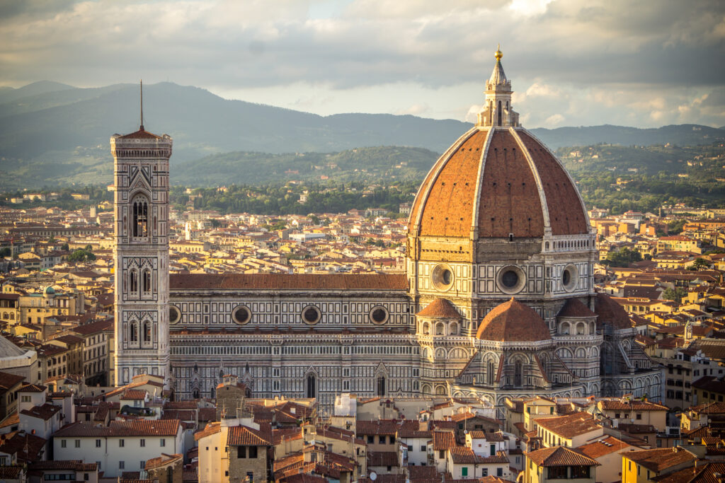 Die imposante Kathedrale Santa Maria del Fiore in Florenz, links hat sie einen Kirchturm, rechts des Kirchenschiffs ragt die große Kuppel in die Höhe. Hoch über den Dächern der Stadt und im Hintergrund sieht man Hügel und Berge