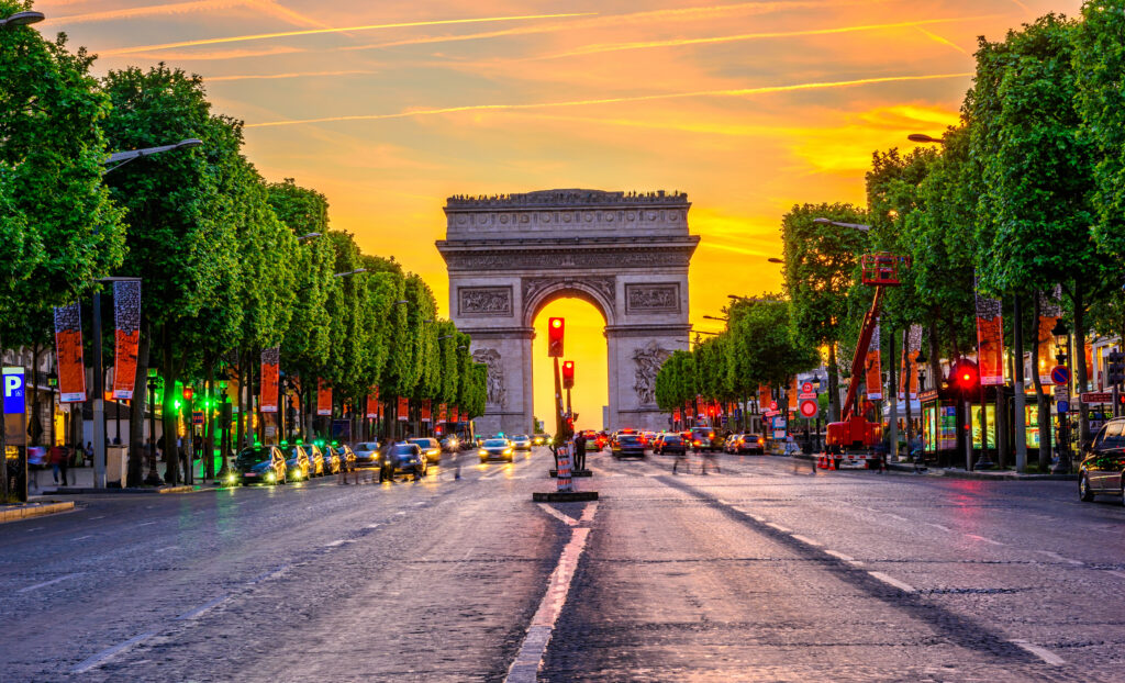 Autos auf der Straße Champs-Élysées, an den Seiten symmetrisch angeordnete Bäume, im Hintergrund ragt der Triumphbogen vor dem orangen Himmel des Sonnenuntergang empor