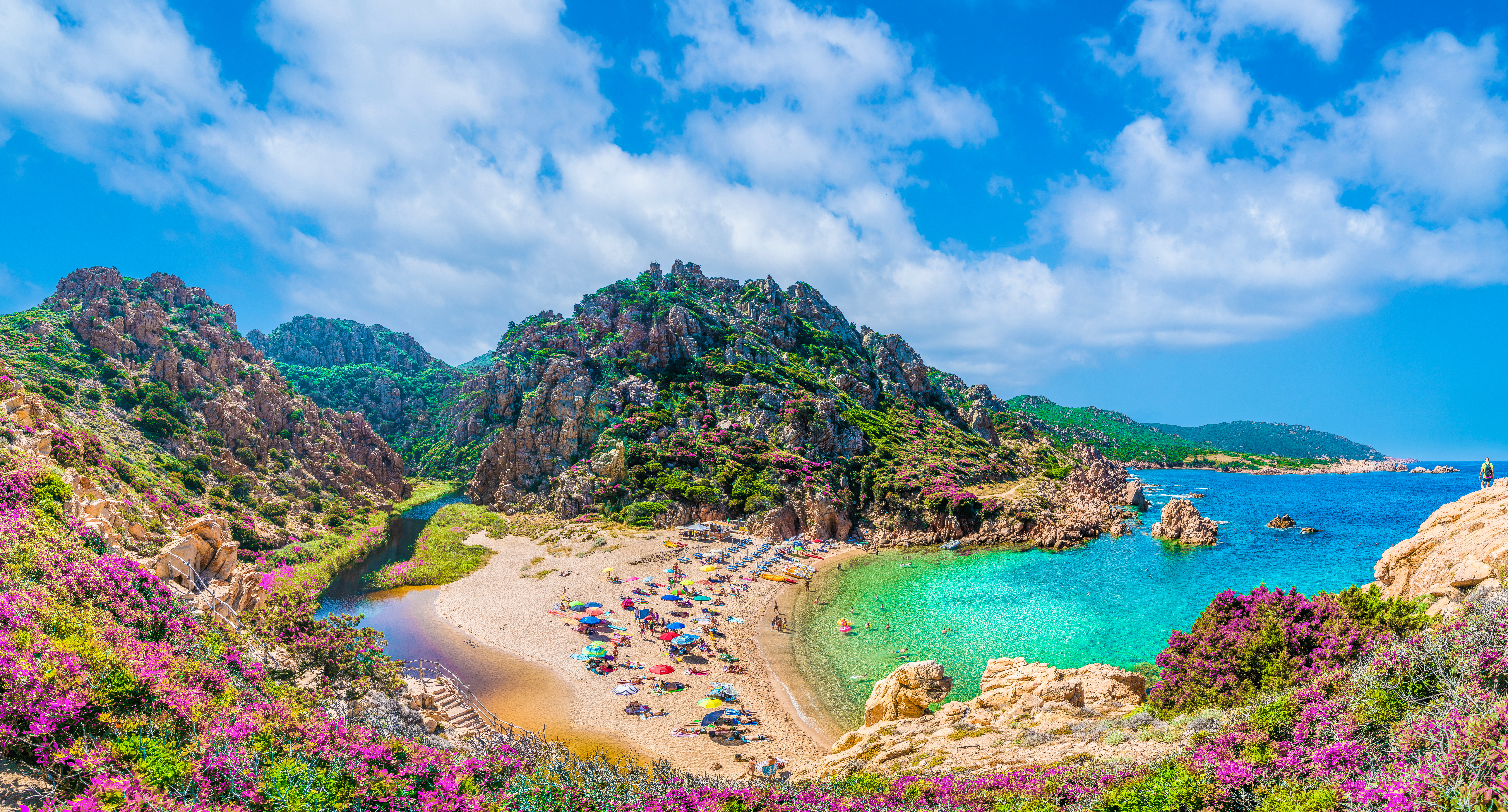 Panoramablick auf einen Strand auf Sardinien, der Strand mit vielen Menschen mit Sonnenschirmen liegt am türkisen Meer, um den Strand herum sind felsige Hügel mit violetten Blumen und Felsformationen