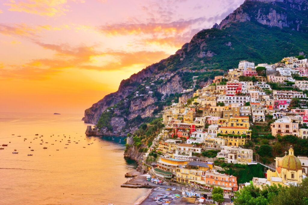 Links das Meer, rechts die bunten Häuser Positanos, die an einem Berghang liegen und bis zu einem Strand reichen. Im Hintergrund der Sonnenuntergang und ein Berg mit Wald und Felsen. 