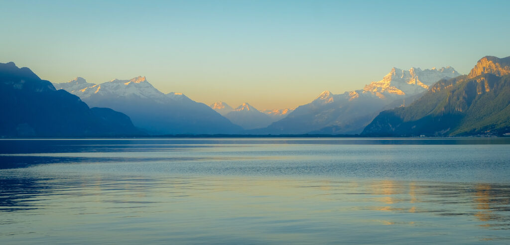 Der Genfer See, im Hintergrund Alpenkulisse mit von der aufgehenden Sonne angeschienenen Berggipfeln 