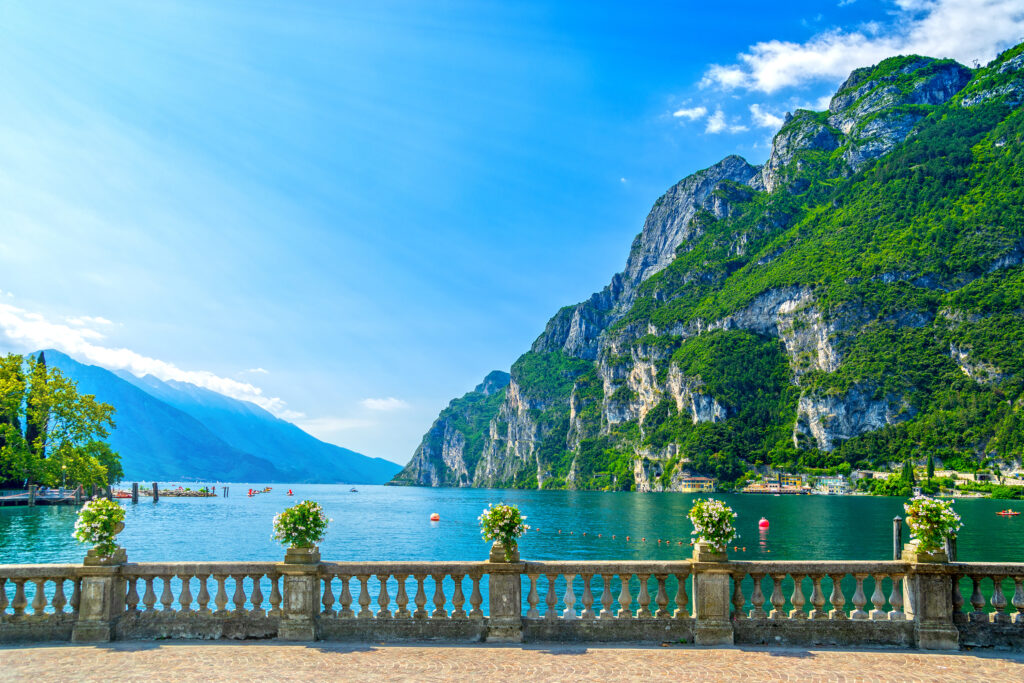 Der Gardasee mit Bergkulisse im Hintergrund, im Vordergrund das Ufer des Sees mit einem Geländer aus Steinsäulen und Blumen