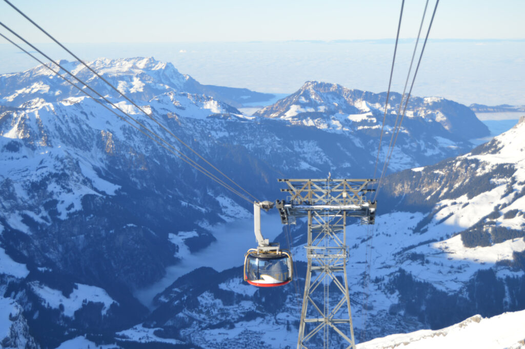 Mittig im Bild die Gondel einer Seilbahn, die auf den schneebedeckten Titlis fährt, weiße Gebirgsketten im Hintergrund