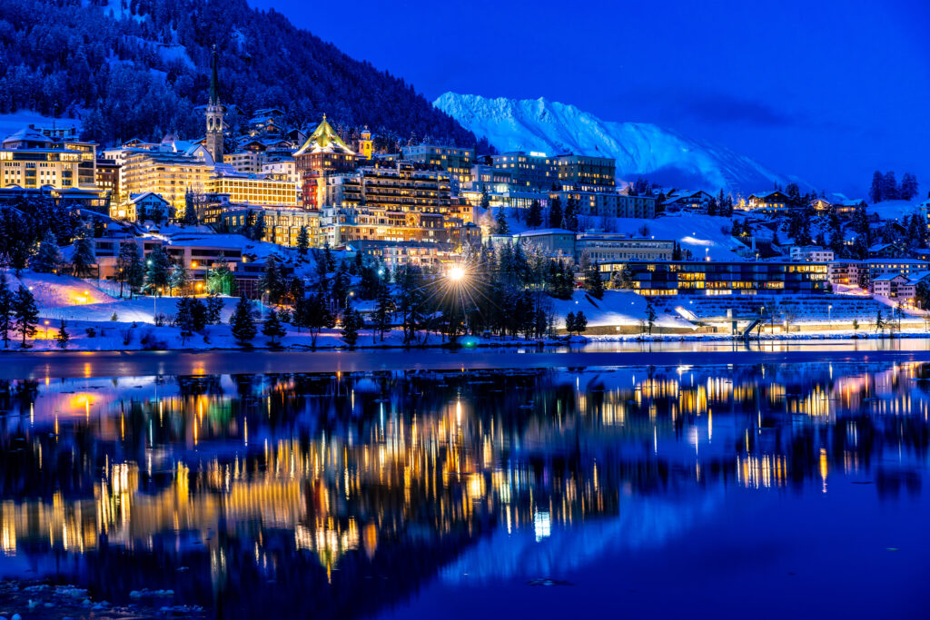 Blick bei Nacht auf das beleuchtete St. Moritz, im Vordergrund der St. Moritz See mit Spiegelungen auf der Wasseroberfläche, im Hintergrund verschneite Berggipfel 