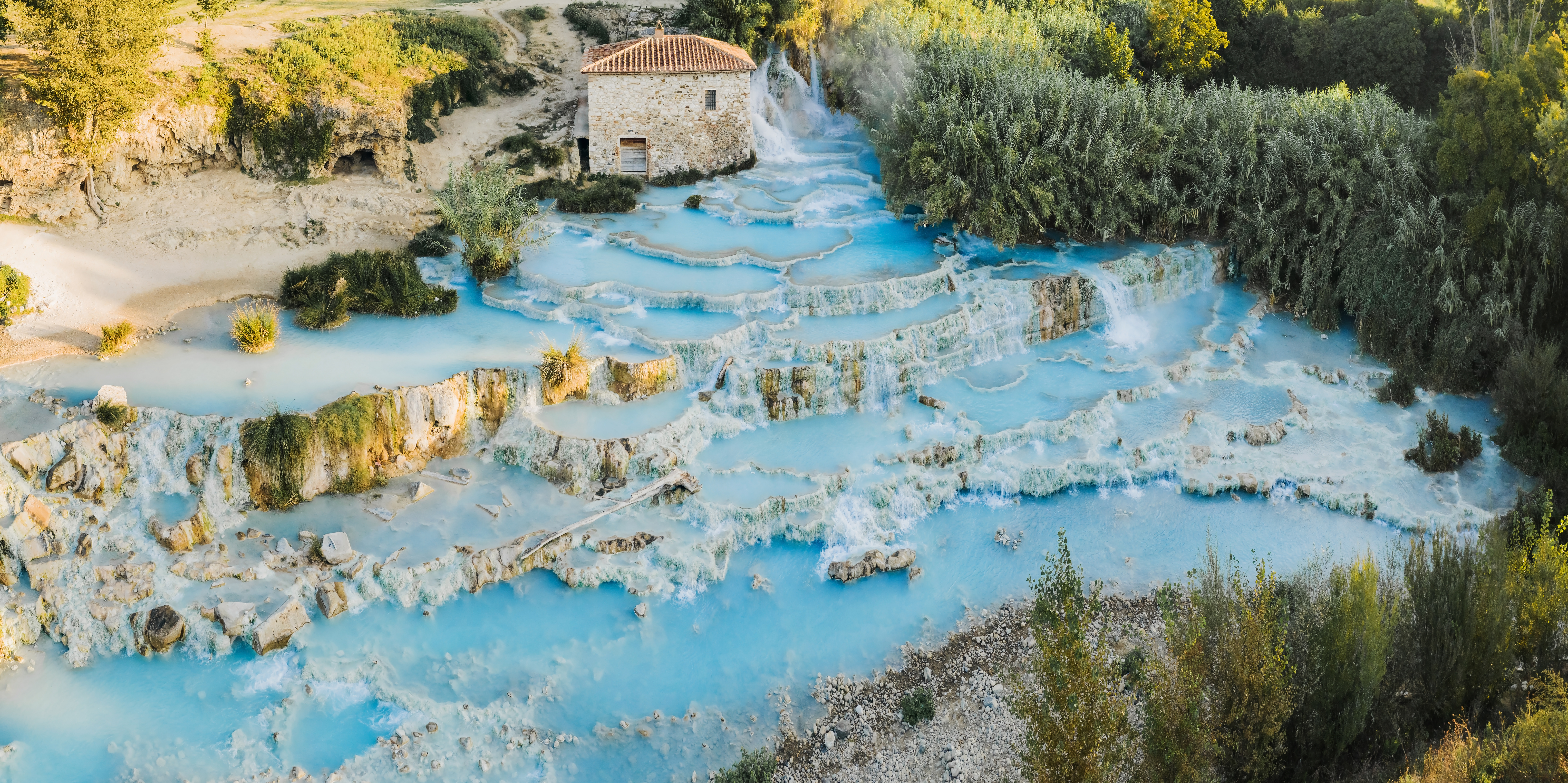 Mittig steht ein beiges Haus, rechts daneben fließt das Wasser der Quellen von Saturnia über einen Wasserfall und verteilt sich auf natürliche Terrassen, das Wasser ist hellblau und dampft