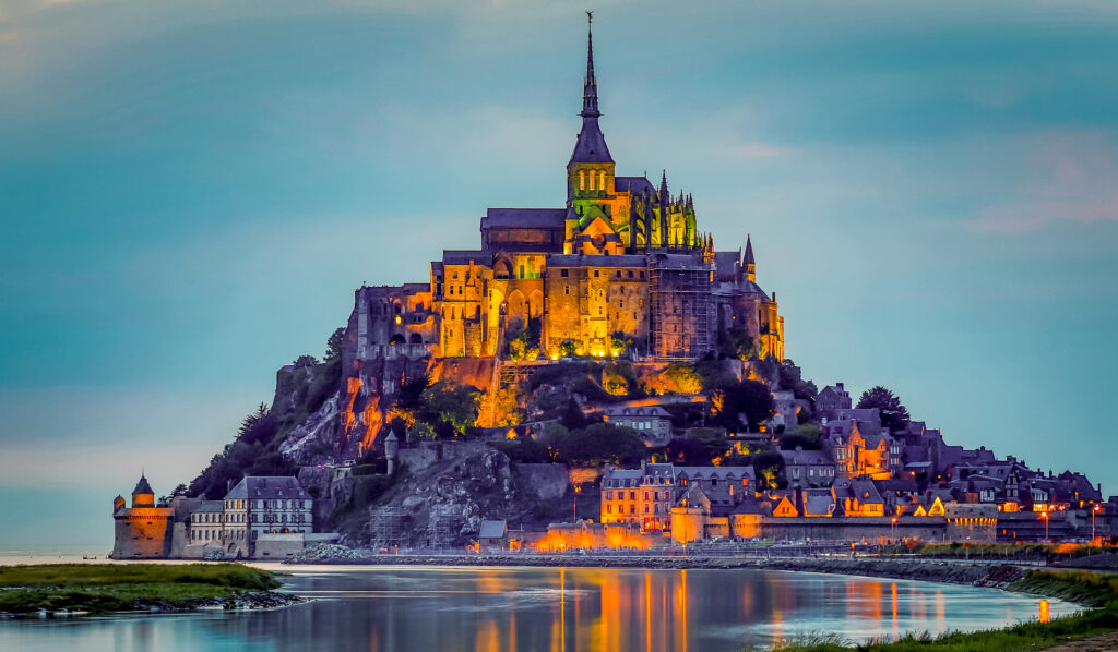 Die beleuchtete Klosterinsel Mont-Saint-Michel in der Dämmerung, umgeben von Wasser, auf der Spitze der Felsinsel liegt die imposante Abtei mit einem hohen Kirchturm