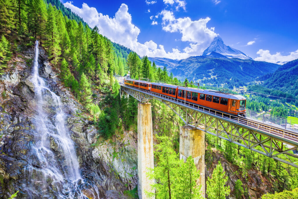 Ein kleiner roter Zug der Gornergrat Bahn fährt über eine Brücke. Links im Bild sieht man einen Wasserfall an einer Felswand und rechts sieht man im Hintergrund die Bergspitze des Matterhorns


























































