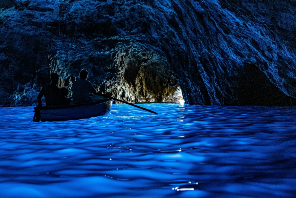 Ein Ruderboot, das in der blauen Grotte in Capri Richtung Ausgang der Höhle treibt. Das Wasser leuchtet in einem schönen Blauton und die Höhle ist ansonsten dunkel.