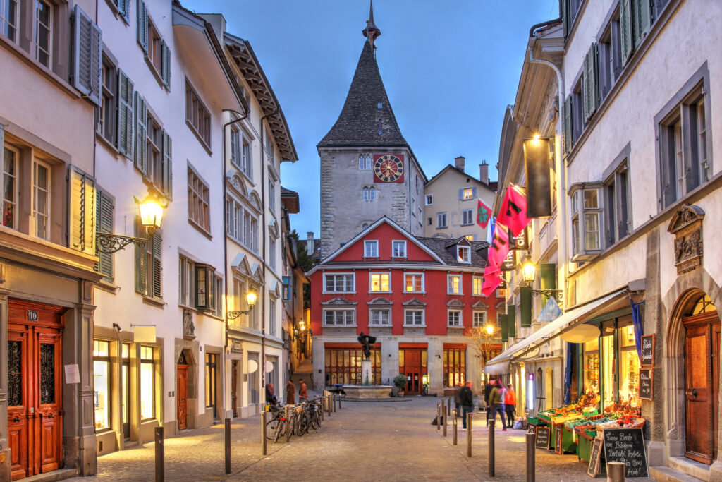 Eine Straße in der Züricher Altstadt, Häuser mit Fensterläden, am Ende der Straße ist ein altes Gebäude mit einem Turm und einer Turmuhr