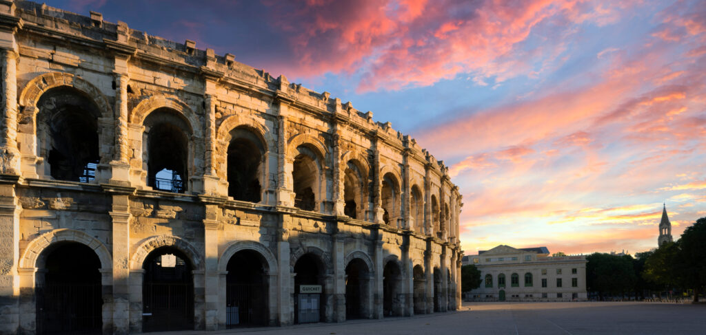 Das Amphitheater von Nîmes mit den bogenförmigen Arkaden, es wird teilweise von der Sonne angeschienen und die Wolken am Himmel sind rosa verfärbt vom Sonnenuntergang