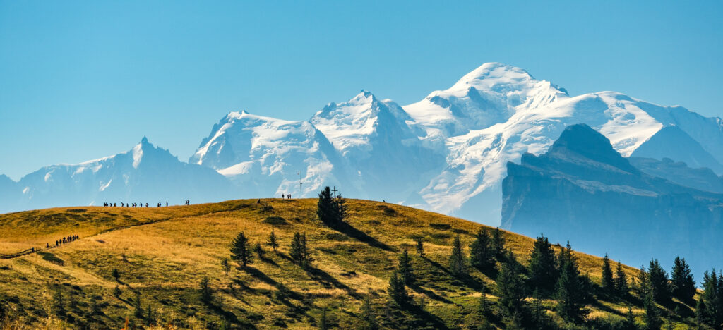 Die weiße Bergspitze des Mont Blancs am Hintergrund, im Vordergrund sieht man die Silhouetten von Wanderern auf einem grünen Hügel