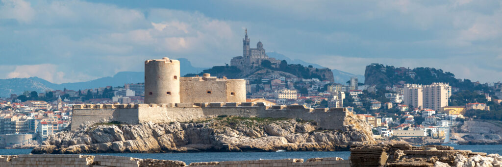 Die Festung Château d'If auf einer Felsinsel im Meer, im Hintergrund die Stadt Marseille mit einer Kirche auf dem Berg