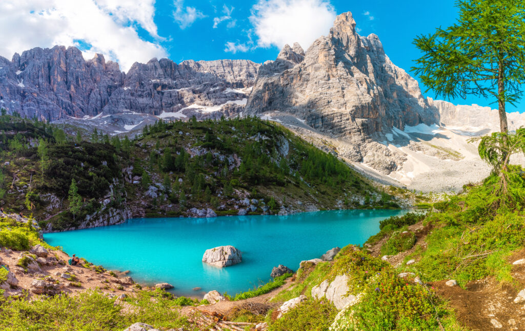 Ein türkisblauer See vor den Felsgipfeln der Dolomiten, Reste von Schnee auf den Felsen, der See ist von Gräsern umgeben