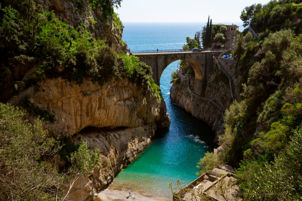 Das türkisblaue Wasser schlängelt sich im Fjord Furore ins Landesinnere zwischen Felsklippen hindurch, am Ende liegt ein kleiner Strand und über dem Fjord reicht eine Brücke von links nach rechts