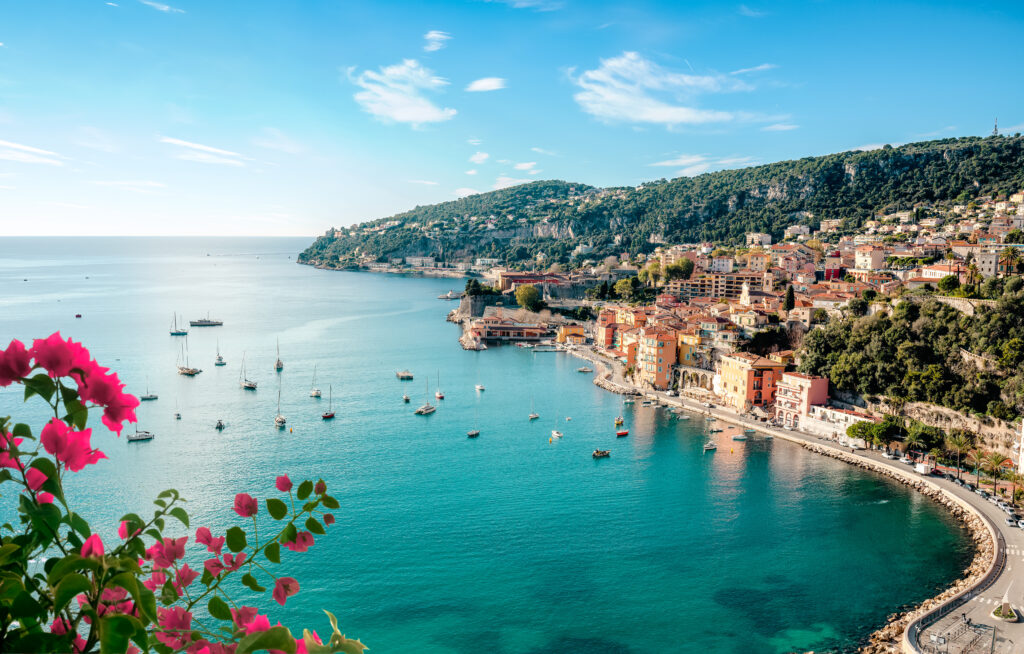 Küstenabschnitt an der Côte d'Azur, links im Bild sind rosafarbene Blüten zu sehen, in der hügeligen Landschaft am Mittelmeer liegt eine kleine Stadt 
