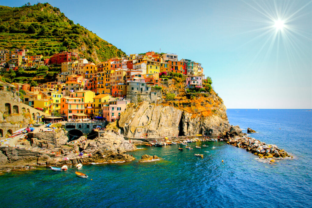 Eines der Dörfer der Cinque Terre Region, bunte Häuser säumen einen Felsvorsprung, im Hintergrund ragen Weinberge in die Höhe, Menschen schwimmen im Wasser und Boote treiben am Ufer im Wasser