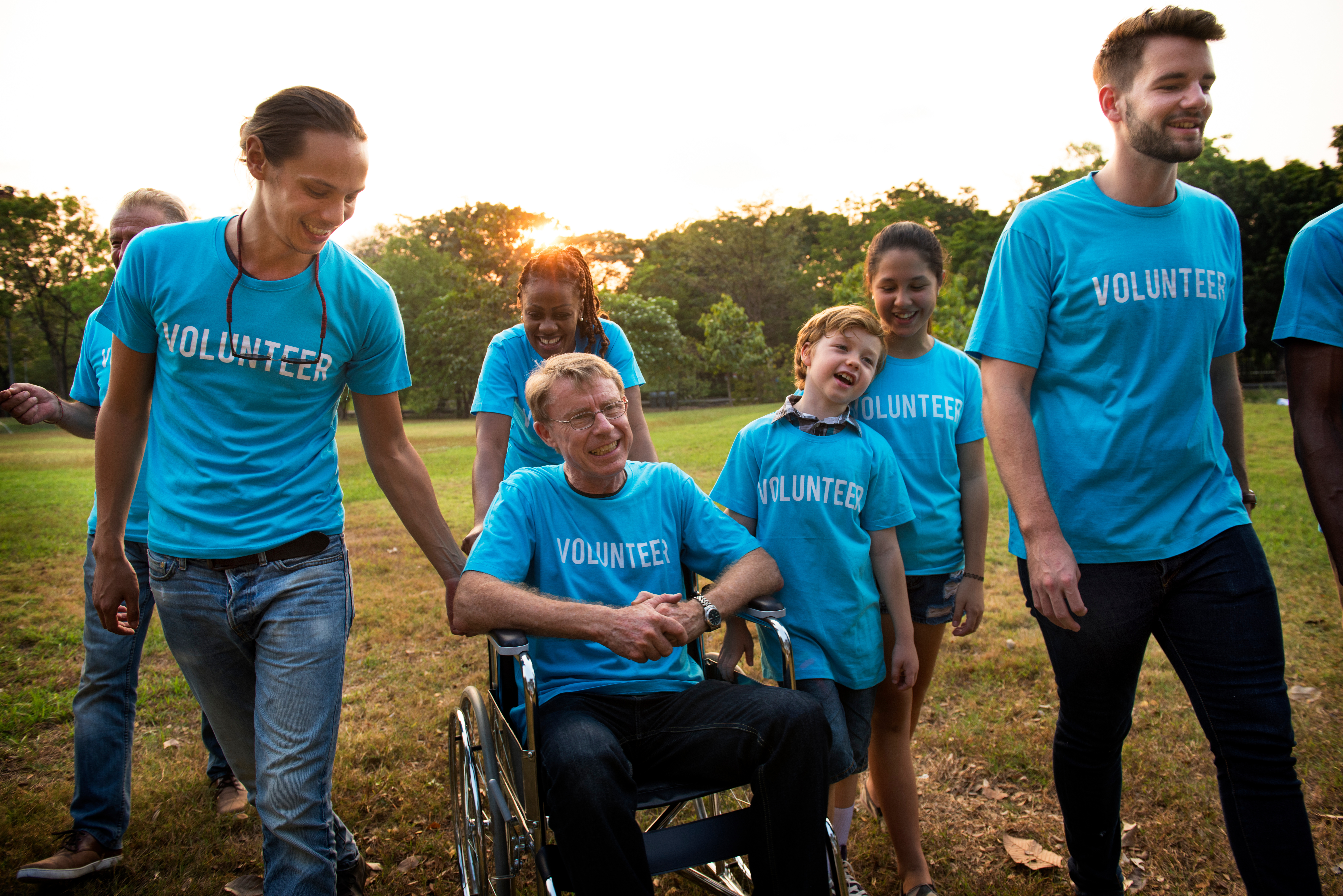 Eine Gruppe von freiwilligen Helfern, die gemeinsam auf einer Wiese stehen, eine Person ist im Rollstuhl. Alle tragen ein blaues Shirt, auf dem "volunteer" steht