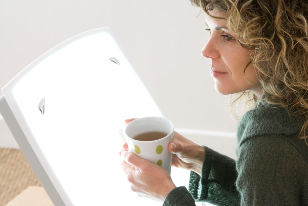 Eine Frau sitzt mit einer Tasse Tee in den Händen vor einer hellen Tageslichtlampe