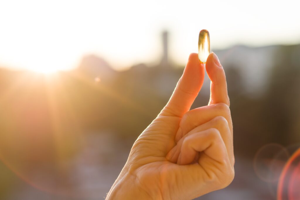 Eine Person hält eine durchsichtige Vitamin D Kapsel in die Luft, Sonnenstrahlen scheinen durch die Tablette