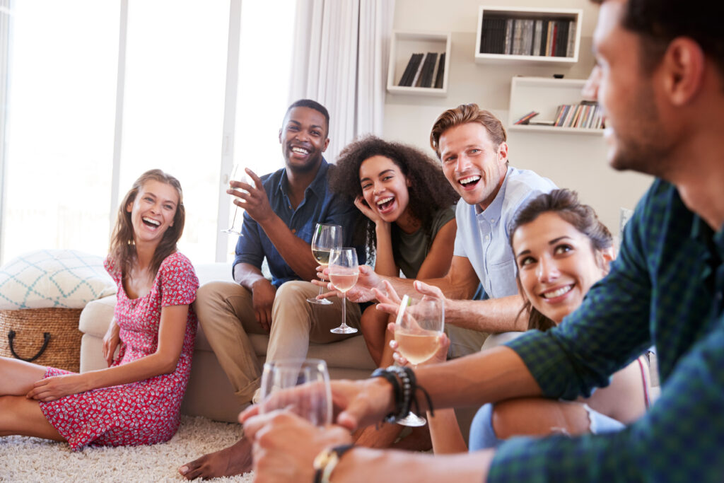 Eine Gruppe von Freunden und Freundinnen sitzt mit Weingläsern und glücklich auf dem Teppichboden und dem Sofa in einem Wohnzimmer