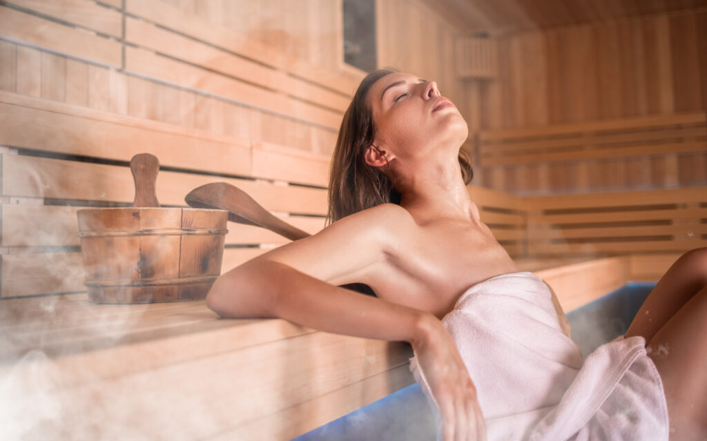 Eine Frau ist in ein Handtuch gewickelt und sitzt entspannt in der dampfenden Sauna