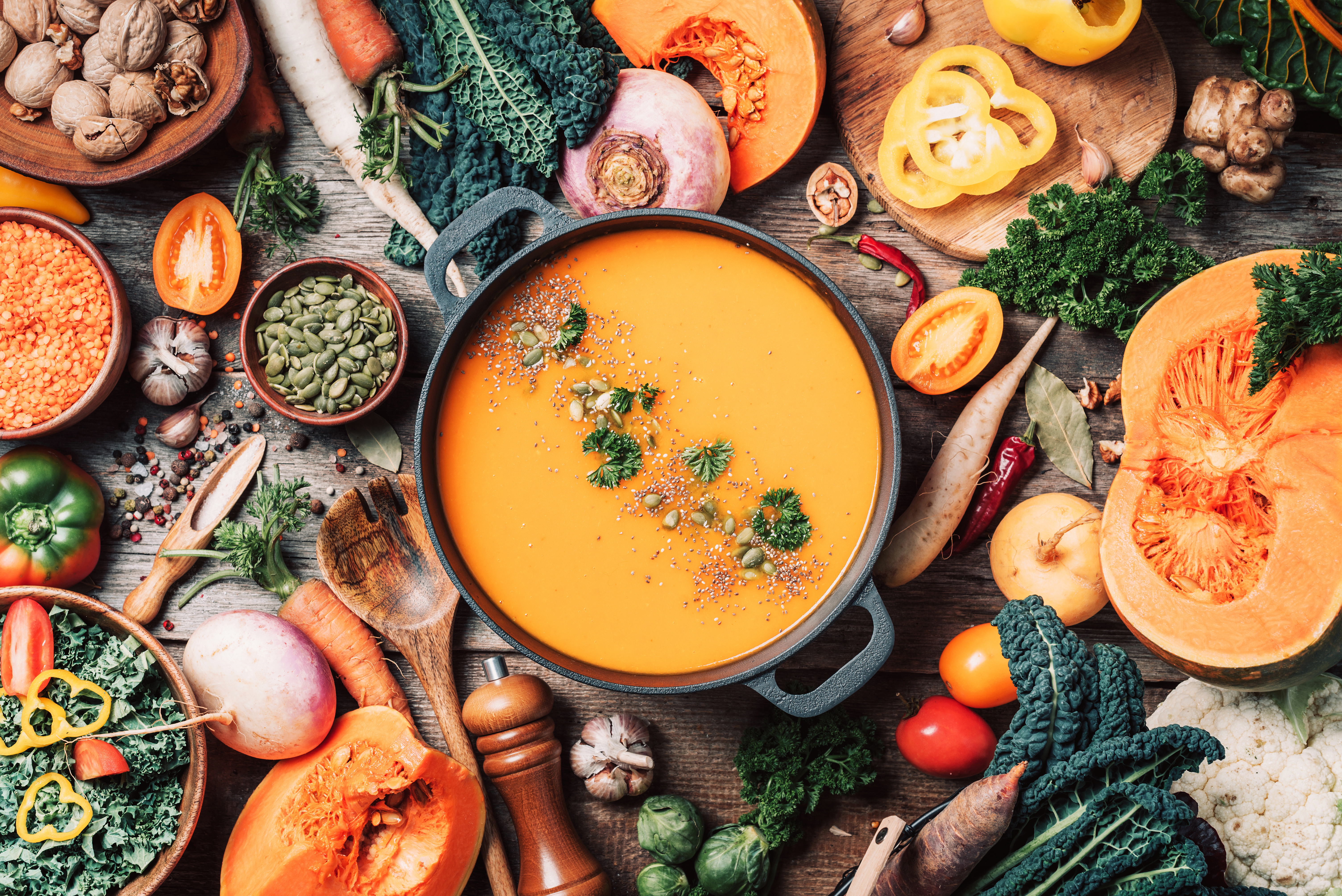 Herbstliches Gemüse: verschiedene Kohlsorten, Karotten, Paprika, Linsen, Kürbis. In der Mitte ist ein Topf mit einer Suppe zu sehen und das Gemüse liegt darum herum auf dem Tisch
