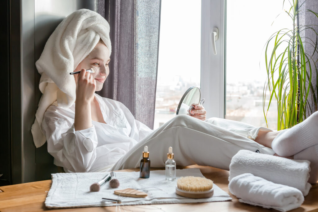 Eine Frau hat einen Handtuchturban auf, trägt mit einem Pinsel eine Gesichtsmaske auf, sie sitzt am Fenster und hat die Füße hochgelegt auf den Tisch. 