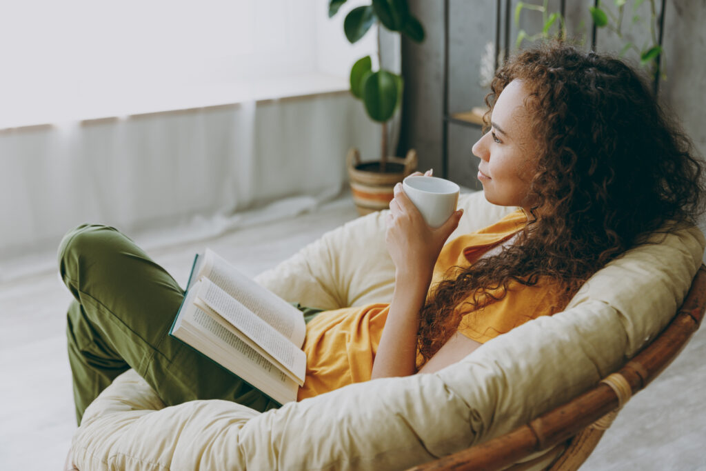 Eine Frau sitzt mit einer Tasse in den Händen und einem Buch auf dem Schoss auf einem gemütlichen Sesses